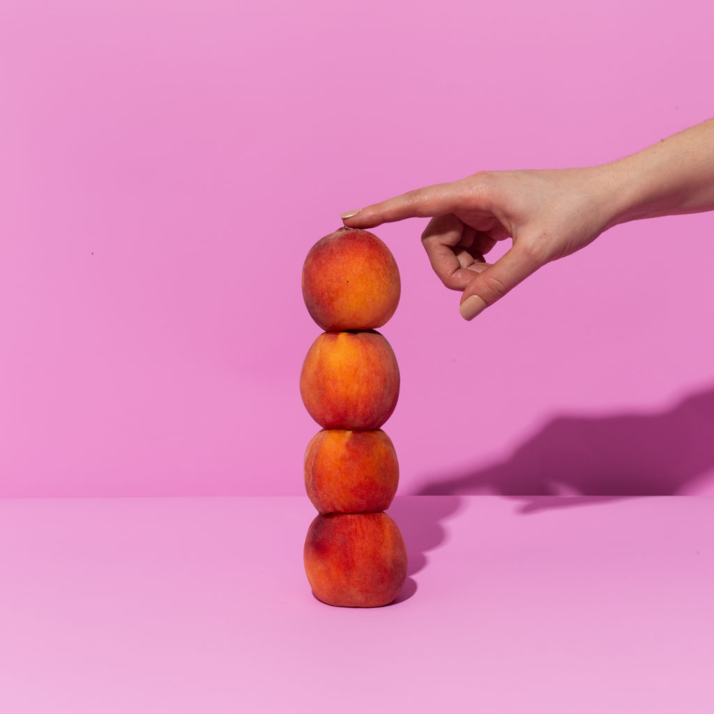 finger reaching out and tapping stack of 4 peaches on a pink background