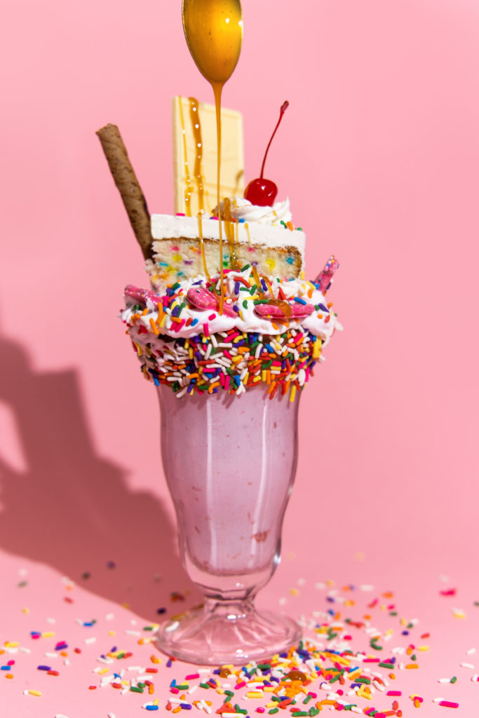 photo of caramel drizzling onto pink milkshake that is topped with rainbow sprinkles, animal crackers, chocolate, and a piece of cake