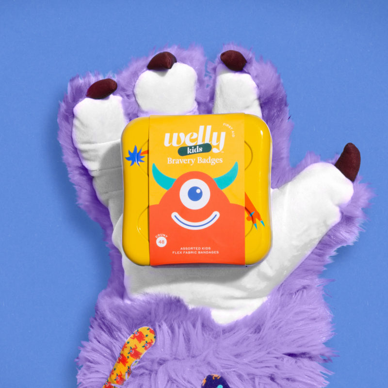 PURPLE MONSTER HAND HOLDING WELLY KIDS BRAVERY BANDAGES ON BLUE BACKGROUND